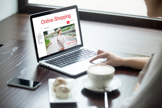 Young woman sitting in cafe with cup of coffee and cakes, working on laptop computer, shopping using online web service. Attractive female browsing shopping website. Closeup view over the shoulder
