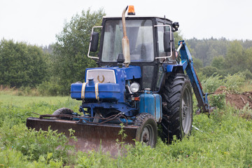 Old blue tractor-excavator stands in a meadow in the rain