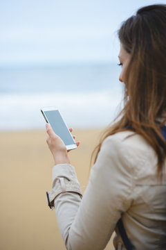 Woman messaging or video calling on smartphone towards the sea during autumn trip to the beach.