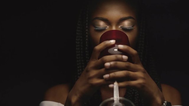 Young beautiful african american female fashion model drinking from red wineglass under lamp in dark