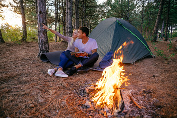 Young caucasian couple camping in the forest. Hikers in the wood near the tent with campfire, ukulele guitar, map and compass. 