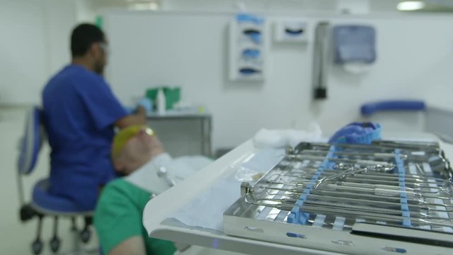 Slight camera track to dentist tray of instruments with dentist and patient in background. 
(Filmed in ProRes with high dynamic range for flexibility for image grading)