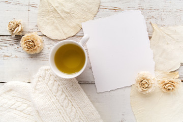 Cup of tea with gloves, flowers and blank card on white wooden background