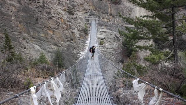 Female trekker walks along suspending bridge with old praying flags flattering in strong wind. She stops in the middle and looks into the distance