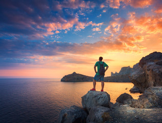 Beautiful summer landscape with standing man with backpack on the stone at the ocean against the colorful sky with clouds at sunset. Travel background. Sport, lifestyle. Tourism