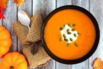 Pumpkin soup topped with pumpkin seeds and cream, above scene on rustic wood background with autumn leaves