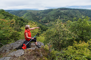 Seniorin sitzt auf einem Felsvorsprung und zeigt mit den Finger; Denntal, Eifel 