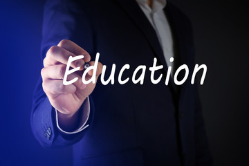 Businessman in suit writing word EDUCATION at transparent whiteboard on dark background
