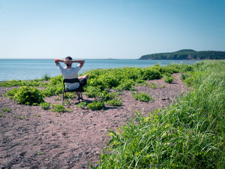 Man sitting on chair on stony beach overlooking a bay with hills