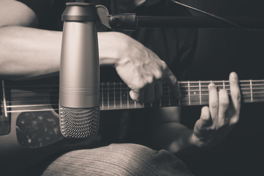 male musician playing acoustic guitar behind condenser microphone in recording studio, bw film filter