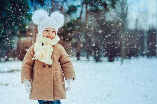 Cute Baby Girl Enjoying Winter Walk In Snowy Park, Wearing Warm Hat And Coat