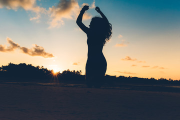 Beautiful silhouette portrait of summer girl jumping with hands in air on white sand in exotic island at sunset.