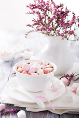 Obraz na płótnie Canvas Cup of hot chocolate with mini marshmallows with pink flowers in jug on white wooden background with copy space.