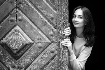 Obraz premium Pretty young girl peering from behind ancient wooden door, black-and-white photo.