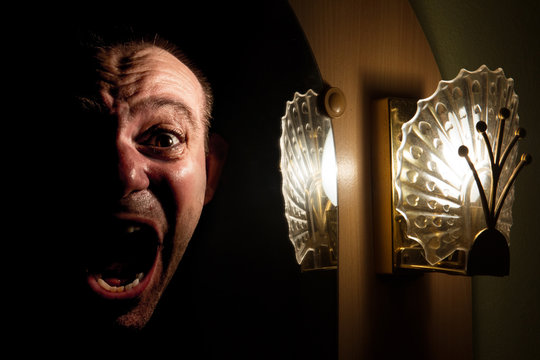 The face of a screaming man in the mirror at night. Halloween haunting. Horrifying scream ghost in the mirror. Poltergeist appears from the shadows and looks from the lighted mirror.