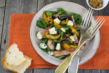 Warm autumn salad with potatoes, mushrooms, bacon, spinach, mustard sauce and cheese on orange napkin. Wooden old background. Selective focus.