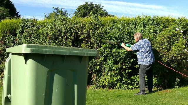Dolly shot of a man trimming a garden hedge in front of a wheely bin.