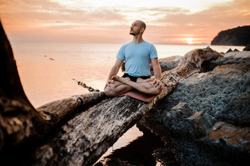 Man meditating on broken tree near sea  sunset . Doing yoga outdoors