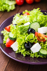 Greek salad (lettuce, tomatoes, feta cheese, cucumbers, black olives) on dark wooden background close up. Healthy food.
