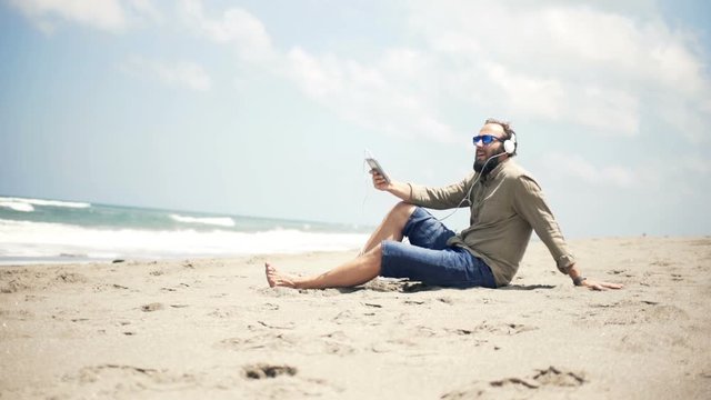 Young man listen to music on cellphone sitting on beach near sea
