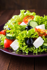 Greek salad (lettuce, tomatoes, feta cheese, cucumbers, black olives) on dark wooden background close up. Healthy food.