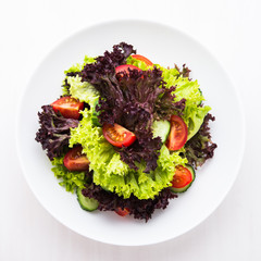 Fresh salad with green and purple lettuce, tomatoes and cucumbers on white wooden background top view. Healthy food.