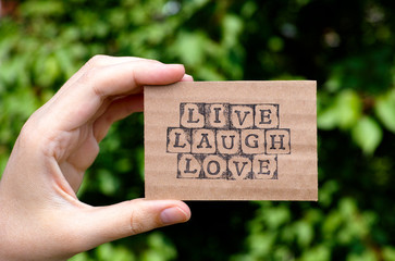 Woman hand holding cardboard card with words Live Laugh Love made by black alphabet stamps opposite green floral background. 