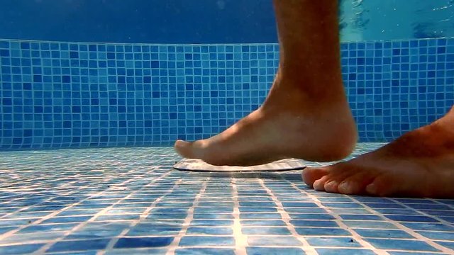 A male feet walking in a swimming pool from right to left with a white strainer in background (underwater). Slow motion.