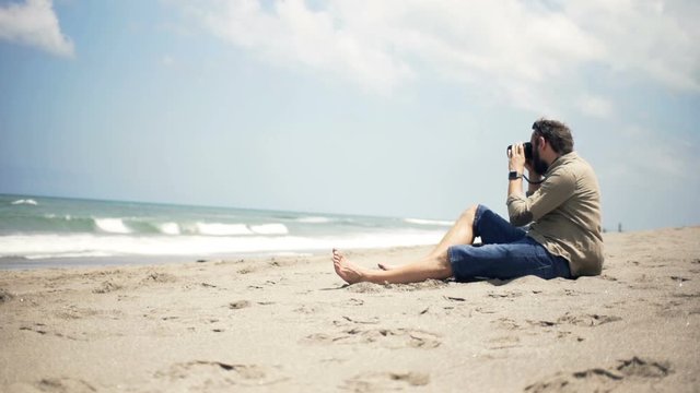 Man taking photo of kite surfer with digital camera sitting on beach 
