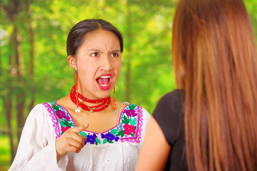 Two beautiful young women posing for camera, one wearing traditional andean clothing, the other in casual clothes, interacting argument, park background