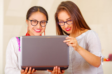 Two beautiful young women posing for camera, one wearing traditional andean clothing, the other in casual clothes, holding laptop between them interacting looking at screen, both smiling, park