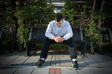 Tired young athlete is resting after workout in a park. He is sitting on the bench and leaning over...