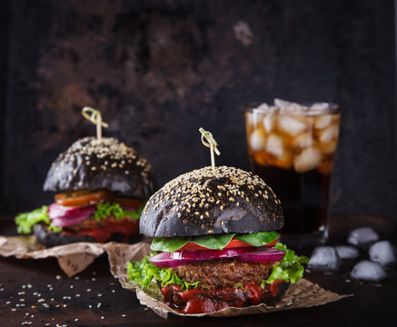 Beef burger with a black bun,with lettuce  served on pieces of brown paper on a rustic wooden table of counter, on a dark background.