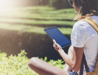 Hipster young girl with backpack using tablet computer, hands holding gadget and texting message on device. View tourist traveler with sunglasses on background flare and bokeh, mockup copy space