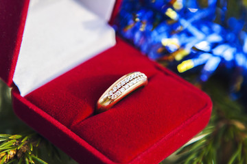 Золотое кольцо в красной коробочке на фоне зеленой елки и синей гирлянды. © Valeriia