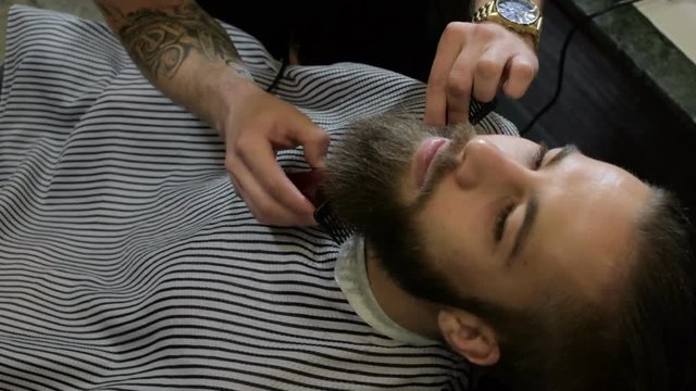 Hairdresser with tattoo serving client in barbershop, making beard haircut using shaver