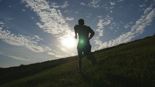 Young men running over green hill over blue sky background. Male athletes is jogging in nature at sunset. Sport runners jogging uphill outdoor at sunrise with flare. Cross-country training. Slowmotion