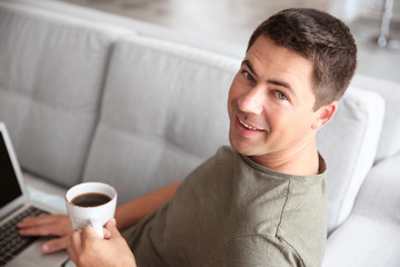 Handsome man sitting on couch with laptop and cup of coffee