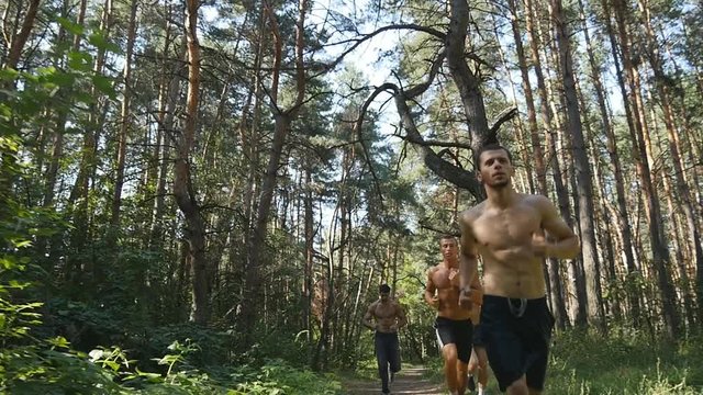 Group of young muscular athletes running at the forest path. Active strong men training outdoors. Fit handsome athletic male sportsmens working out. Sport men running through the woods. Slow motion