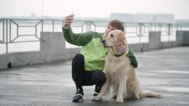 Lockdown wide shot of teenage boy in windcheater sitting on embankment with his golden retriever dog petting him and taking selfie