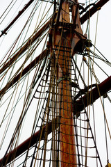 Fototapeta premium The wooden mast and lots of ropes on a large sailboat. Gray sky above.