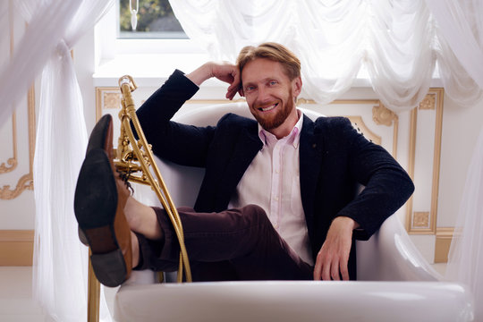 Picture of happy smiling rich blond man in expensive apartments. Cheerful young man lying in bath and looking at camera.