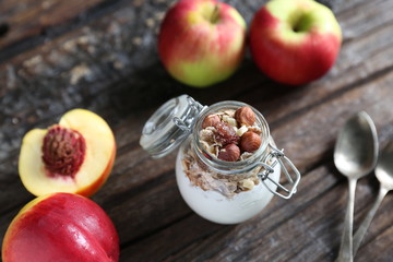 Glass jar with yogurt, granolla and almond nuts in it. Nectarin and apples are on the wooden backgound with spoons closed to the jar.