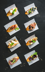 Italian crostini with various toppings on white baking paper over black plywood background, top view