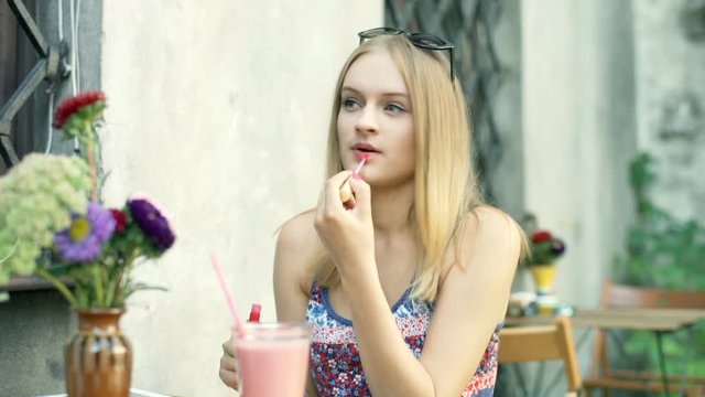Pretty, blonde girl using lipgloss while painting her lips in the outdoor cafe
