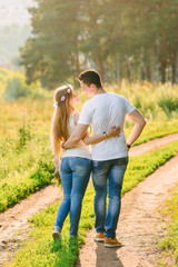 A nice couple in blue jeans and white t-shirts looking at each other happily standing back to the camera green grass and trees all around their path