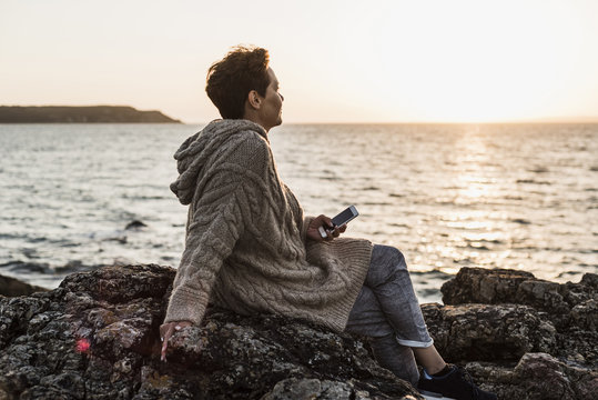 France, Crozon peninsula, woman with smartphone sitting on rocks at sunset