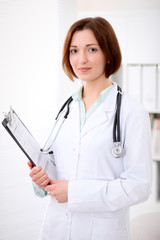 Young brunette female doctor standing with clipboard and smiling at hospital office. Health care, insurance and help concept. Physician ready to examine patient