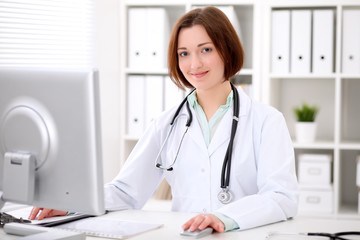 Young brunette female doctor sitting at a desk and working on the computer at the hospital office.  Health care, insurance and help concept. Physician ready to examine patient