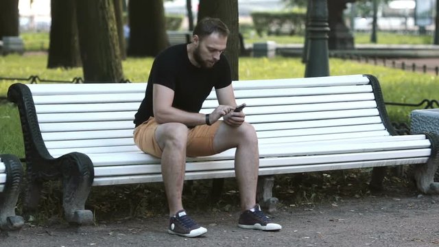 smiling attractive young man sitting on a bench in the park and using a smartphone